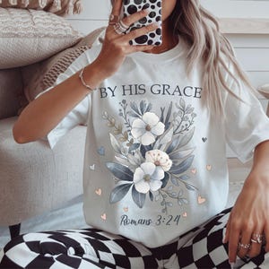 May include: White t-shirt featuring a floral design with white and light blue flowers, green leaves, and the text "BY HIS GRACE" and "Romans 3:24". The design includes small pink and blue hearts.
