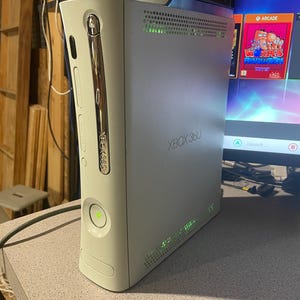 May include: A white Xbox 360 console with a silver trim and the Xbox 360 logo. The console has a power button and a disc tray. The console is on a gray surface.