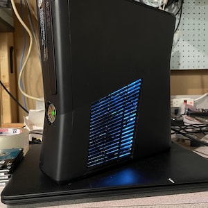 May include: Black Xbox 360 console with a vertical design. The console features a vent with blue internal lighting and the Xbox 360 logo. A green light is visible on the front. The console is on a black base.