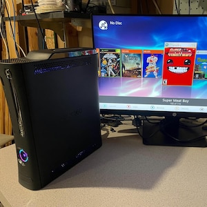 May include: Black Xbox 360 console with the Xbox 360 logo. A monitor displays a game menu with the text "No Disc" and game titles like "Super Meat Boy". The console has blue and purple lights.