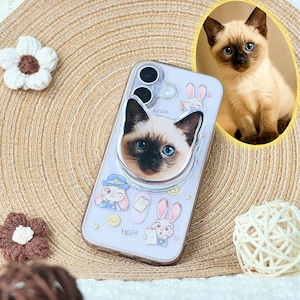 Può includere: Una cover trasparente per smartphone con un supporto circolare estraibile raffigurante il muso di un gattino siamese. La cover è decorata con personaggi di coniglietti, piccole stelle, faccine sorridenti e la scritta "HOP!" stampata.