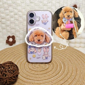 Può includere: Una cover trasparente per smartphone con illustrazioni di conigli e il testo HOP!, dotata di un supporto circolare con la foto di un barboncino marrone. Un riquadro circolare mostra lo stesso barboncino con una pettorina blu e un accessorio rosa.