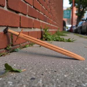 Puede incluir: Una espada de madera con acabado marrón claro descansa sobre una superficie de hormigón junto a una pared de ladrillo rojo. La espada tiene una guarda y una empuñadura detallada. La escena es al aire libre, con vegetación y una calle visible al fondo.