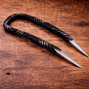 Può includere: Una forcina per capelli in metallo nero forgiata a mano con un design a spirale. Le due punte sono affilate. L'accessorio per capelli è esposto su una superficie di legno.