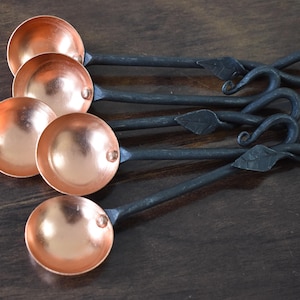 Könnte beinhalten: Ein Set aus fünf handgefertigten Messlöffeln aus Kupfer mit dunklen Metallgriffen. Die Kupferschalen haben eine polierte Oberfläche, und die Griffe weisen dekorative Blatt- und Wirbelmuster auf. Die Löffel sind auf einer dunklen Holzoberfläche angeordnet.