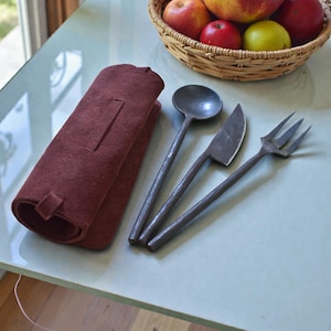 Könnte beinhalten: Ein handgefertigtes Besteckset aus dunklem Metall, bestehend aus einem Löffel, einem Messer und einer Gabel. Das Besteck liegt neben einer aufgerollten, weinroten Leder-Utensilientasche. Im Hintergrund ist ein Korb mit Äpfeln zu sehen.