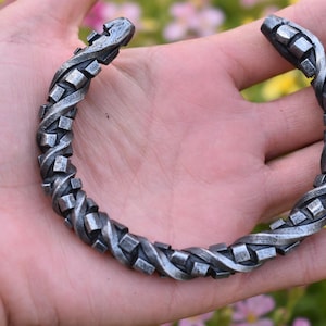 Puede incluir: Una pulsera de metal plateado con un diseño retorcido y texturizado. La pulsera es abierta y presenta una serie de eslabones cuadrados entrelazados. La pulsera tiene aproximadamente 18 cm de circunferencia.