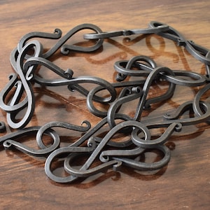 May include: A handcrafted, dark grey metal chain with a series of interconnected, stylised links. Each link features a looped design, creating a decorative and artistic appearance. The chain is displayed on a wooden surface.
