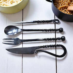 May include: Hand-forged black iron cutlery set. Includes a spoon, fork, knife, and a pointed utensil. Each piece features a twisted handle design. The utensils are arranged on a white wooden surface, with a bowl of pasta and a pot of noodles in the background.