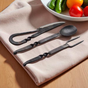 May include: Hand-forged black metal cutlery set including a knife, spoon, and fork. Each piece features a twisted handle design. The set is displayed on a beige cloth, with a bowl of fresh vegetables in the background.