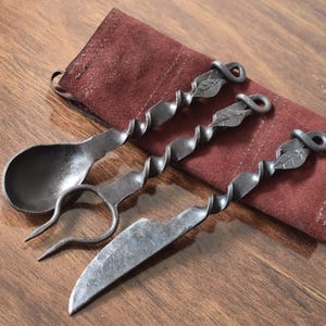 May include: Hand-forged iron flatware set including a spoon, fork, and knife, with twisted handles and leaf accents. The set rests on a burgundy leather pouch, ideal for rustic dining or gifting.