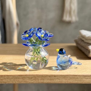 May include: A clear glass spherical vase containing a bouquet of small blue glass flowers with yellow centers, accompanied by a small glass bird figurine featuring blue and white coloring with a yellow beak, both resting on a wooden surface.