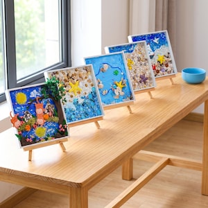 以下が含まれることがあります： 小さな木製のイーゼルに飾られた、白いフレームの正方形のアート作品4点。各フレームには、小さな貝殻、小石、ガラスビーズ、彫刻されたフィギュアなど、さまざまな素材を使用した立体的な風景が収められています。