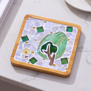 May include: A square coaster with a light wood frame featuring a mosaic design. The surface contains white and translucent glass pieces, small green square tiles, a yellow flower shape, and a central green marbled oval with a brown tree graphic.