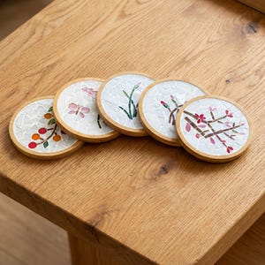 May include: A set of five circular coasters with light wood frames and white mosaic tile centers. Each coaster features a unique nature-inspired design, including orange berries, a pink butterfly, green grass, flower buds, and pink blossoms.