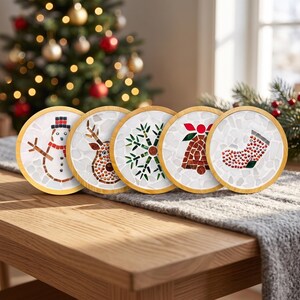 Può includere: Un set di cinque sottobicchieri circolari con cornici in legno chiaro, ognuno caratterizzato da un design a mosaico unico su sfondo bianco. I disegni includono un pupazzo di neve, una renna, un fiocco di neve verde, una campana rossa e una calza.