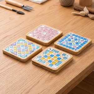 May include: Four square coasters with light wood frames, each featuring a unique mosaic inlay of small glass tiles in various shapes and colors, including shades of blue, yellow, pink, and clear, arranged in geometric patterns on each surface.