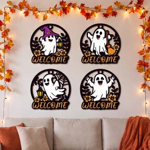 May include: Four circular black wall signs featuring white cartoon ghosts, orange flowers, and the word WELCOME in orange script. One ghost wears a purple hat and holds a lantern, one holds a broom, one holds a lantern, and one has raised arms.