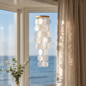 Peut inclure: Un mobile suspendu composé de multiples disques de coquillages blancs circulaires et translucides, fixés à un cadre circulaire en bois clair. Les disques sont disposés en brins verticaux, créant un effet de cascade superposé qui capte la lumière.