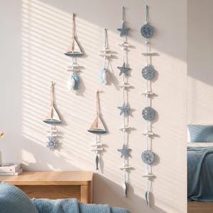 Puede incluir: Un conjunto de seis decoraciones de pared colgantes con motivos náuticos. El set incluye tres adornos de veleros con detalles de peces o ruedas, un adorno de pez pequeño y dos guirnaldas verticales largas con estrellas de mar y formas de dólar de arena.
