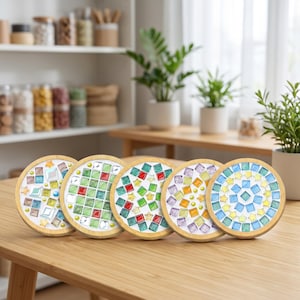 Könnte beinhalten: Fünf runde Untersetzer mit hellem Holzrahmen, die jeweils ein einzigartiges Mosaikmuster aus kleinen, bunten Glasfliesen in Blau-, Grün-, Rot-, Gelb- und Violetttönen aufweisen, ergänzt durch sternförmige Akzente auf einem weißen Untergrund.
