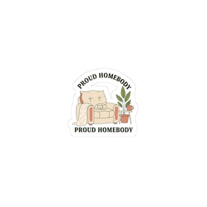 May include: A sticker featuring a beige armchair with a cat, a blanket, a small table with a plant and a mug. The text "PROUD HOMEBODY" is written above and below the illustration in a semi-circle.
