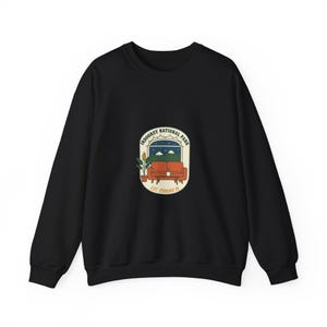 Peut inclure: Sweat-shirt noir avec un motif circulaire représentant une scène de fenêtre. Le motif comprend un canapé rouge, des montagnes et le texte "INDOORSY NATIONAL PARK EST. STAYING IN."