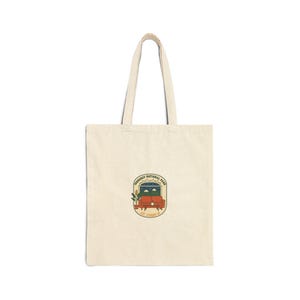 May include: Cream-coloured tote bag with a rectangular patch featuring a red couch, plants, and the text "Dendroast National Park, Est. Standing '76". The bag has two handles and is made of a sturdy fabric.