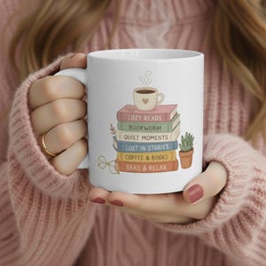 Puede incluir: Taza de cerámica blanca con un diseño de libros apilados, cada uno con un título como "Cozy Reads" y "Bookworm". También se representa una taza de café y una pequeña planta. La taza está sujeta por una persona.