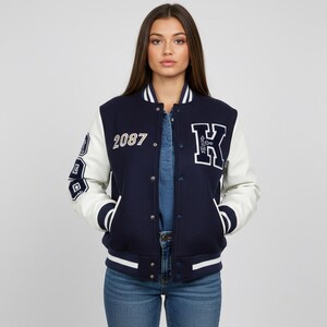 May include: Navy blue and white varsity jacket with the number "2087" and a large "K" patch. The jacket has white sleeves and trim, with a snap-button closure. The jacket is worn over a denim shirt and blue jeans.
