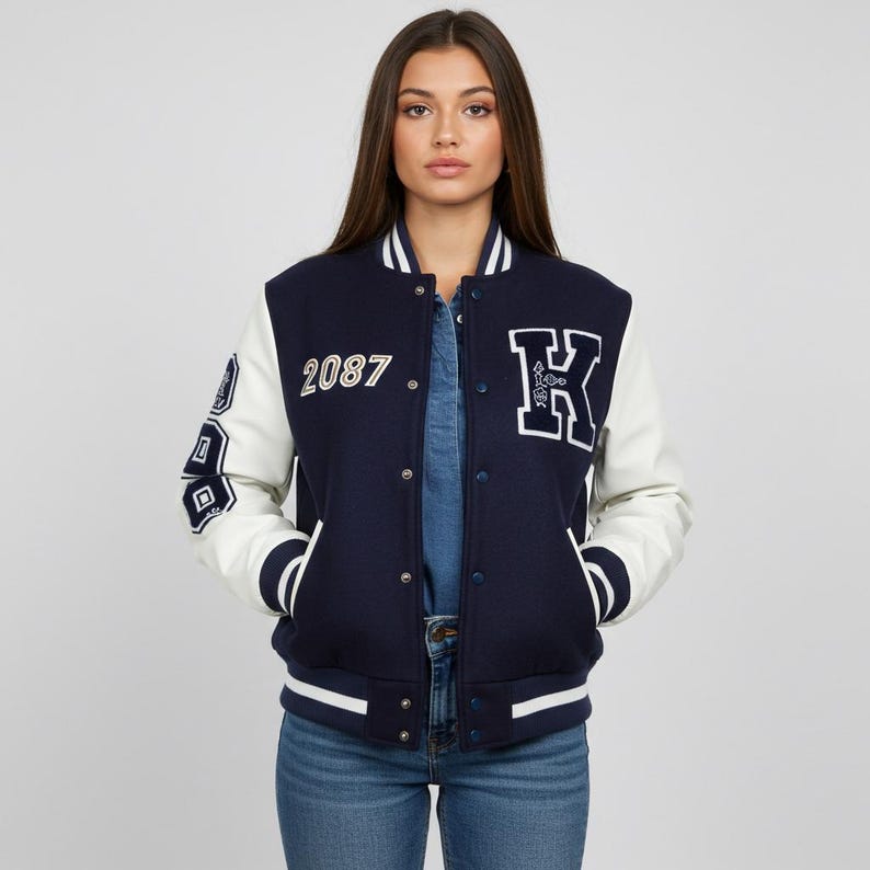 May include: Navy blue and white varsity jacket with the number "2087" and a large "K" patch. The jacket has white sleeves and trim, with a snap-button closure. The jacket is worn over a denim shirt and blue jeans.