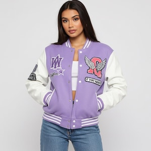 May include: A lavender varsity jacket with white sleeves. The jacket features multiple patches, including a large "Q" with wings and the text "IT YOGE YADU". The jacket has a button-up front and striped detailing.