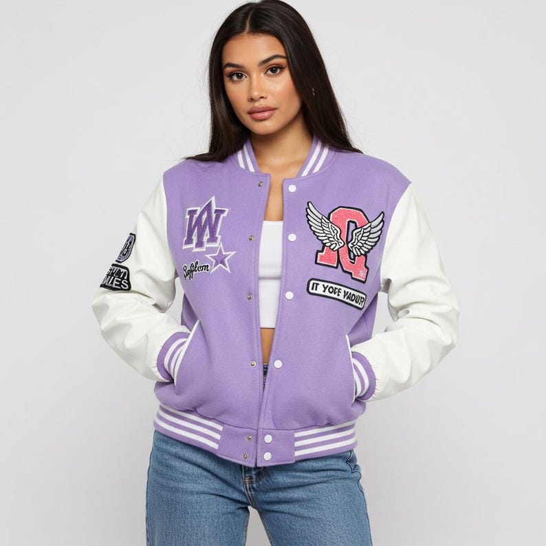 May include: A lavender varsity jacket with white sleeves. The jacket features multiple patches, including a large "Q" with wings and the text "IT YOGE YADU". The jacket has a button-up front and striped detailing.