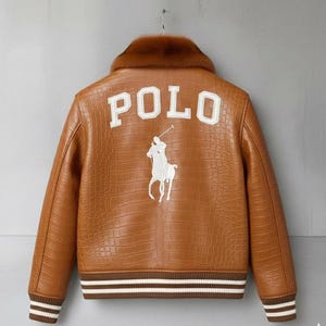 Könnte beinhalten: Braune Lederjacke mit Krokodiltextur, braunem Pelzkragen und weißem "POLO"-Schriftzug auf dem Rücken. Die Jacke hat ein weißes, gesticktes Polospieler-Logo und braun-weiß gestreifte Bündchen.