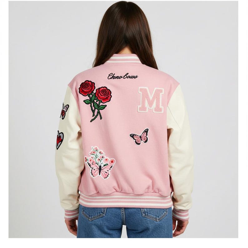 May include: Pink and cream varsity jacket with embroidered roses, butterflies, and a large letter "M". The jacket features the text "Chino Bows" on the back and has cream-colored sleeves and a striped hem.