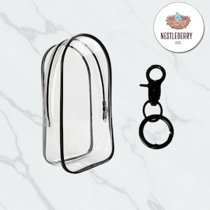 Puede incluir: Una bolsa de cosméticos transparente con cremallera, ribete negro y un llavero de metal negro. La bolsa permite ver el contenido. El logotipo de Nestléberry Co. está en la esquina superior derecha.