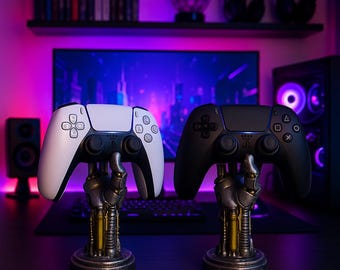 Puede incluir: Dos mandos de juego, uno blanco y otro negro, expuestos sobre soportes decorativos con forma de mano. Los soportes tienen un acabado metálico con detalles dorados. El fondo presenta un monitor de ordenador, altavoces y estanterías con accesorios de juego.
