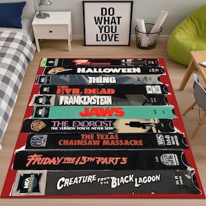 Puede incluir: Una alfombra rectangular con un diseño de cintas VHS apiladas, con títulos de películas como "The Shining" y "Halloween". La alfombra tiene un borde rojo y está colocada sobre un suelo de madera. La frase "Do What You Love" está en un marco negro en el fondo.