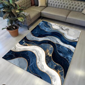 May include: A rectangular rug featuring an abstract design in shades of blue, grey, and white, with gold detailing. The rug is placed on a light wooden floor, near a grey sofa and a potted plant. The rug's design resembles flowing water.