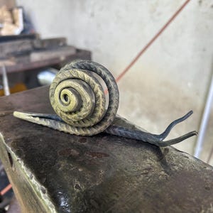 Könnte beinhalten: Eine handgefertigte Metallskulptur einer Schnecke mit spiralförmigem Gehäuse und strukturierter Oberfläche. Die Schnecke besteht aus dunklem Metall mit einem helleren, goldgrünen Farbton auf dem Gehäuse. Sie liegt auf einer dunklen, strukturierten Oberfläche.