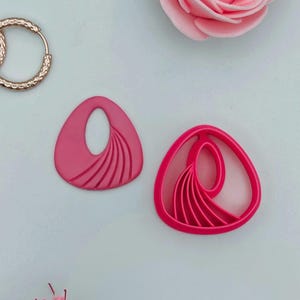 Peut inclure: Deux composants de boucles d'oreilles roses en forme de goutte d'eau avec une découpe ovale et des lignes courbes. Un moule à boucles d'oreilles assorti est à côté du composant de la boucle d'oreille. Une boucle d'oreille dorée et une rose rose sont également dans l'image.