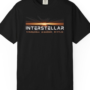 May include: Black t-shirt with the word "INTERSTELLAR" in orange and white block letters. Below the word is the text "1I/OUMUAMUA 2I/BORISOC 3I/ATLAS". The shirt has a graphic of a sunset or sunrise. The shirt is a Comfort Colors brand.