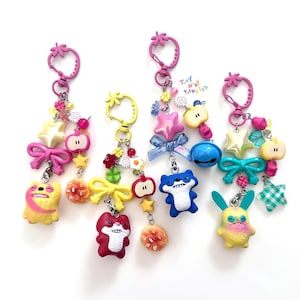 May include: Five vibrant keyrings with assorted charms, such as stars, bows, and cartoon characters. Each keyring features a pink strawberry-shaped clasp. The charms include a yellow sloth, a red fox, a blue fox, and a yellow and pink bunny.