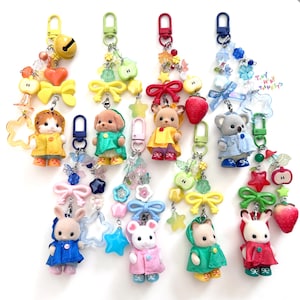 Op de afbeelding: Een verzameling kleurrijke sleutelhangers met miniatuur dierfiguren in verschillende outfits. Elke sleutelhanger bevat decoratieve kralen, strikken en fruitbedels. De sleutelhangers hebben een verscheidenheid aan kleuren, waaronder geel, groen, rood en blauw.