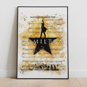 May include: Framed art print featuring a watercolor design with musical notes and the text "ALEXANDER HAMILTON". A black silhouette of a person stands atop a black star with the word "HAMILTON" in white.