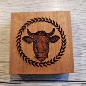 Könnte beinhalten: Quadratische Holzbox mit detaillierter Gravur eines Kuhkopfes in einem Lorbeerkranz. Die Kuh ist braun, der Kranz schwarz, auf hellem Holz. Das Design ist mittig auf der Box platziert.