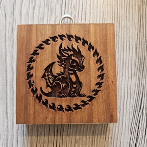 Könnte beinhalten: Quadratischer Holzschmuck mit einem lasergravierten Design eines Drachen in einem kreisförmigen Rahmen aus Flammen. Der Drache ist dunkelbraun, und das Holz hat eine natürliche Maserung. Oben befindet sich eine silberne Metallschlaufe zum Aufhängen.