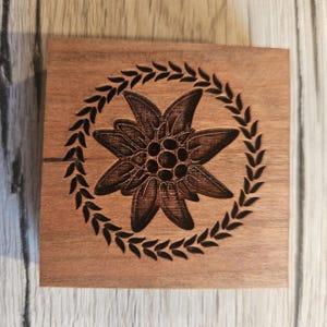 Könnte beinhalten: Eine quadratische Holzbox mit einem geschnitzten Design. Das Design zeigt eine detaillierte Edelweißblume, die von einem Lorbeerkranz umgeben ist. Das Holz hat einen warmen, natürlichen Farbton, und die geschnitzten Elemente sind dunkler.
