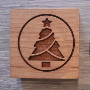 Könnte beinhalten: Quadratischer Holzuntersetzer mit einem lasergeätzten Weihnachtsbaumdesign. Der Baum befindet sich in einem Kreis, der von einem Stern gekrönt wird. Der Untersetzer ist aus hellbraunem Holz gefertigt.