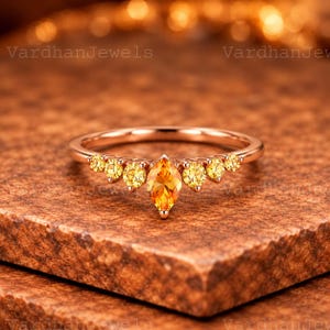 Op de afbeelding: Een ring van roségoud met een centrale oranje edelsteen in marquise-slijpvorm, geflankeerd door drie ronde gele edelstenen aan elke kant. De ringband is dun en gepolijst, met een symmetrische opstelling van zeven stenen in totaal.